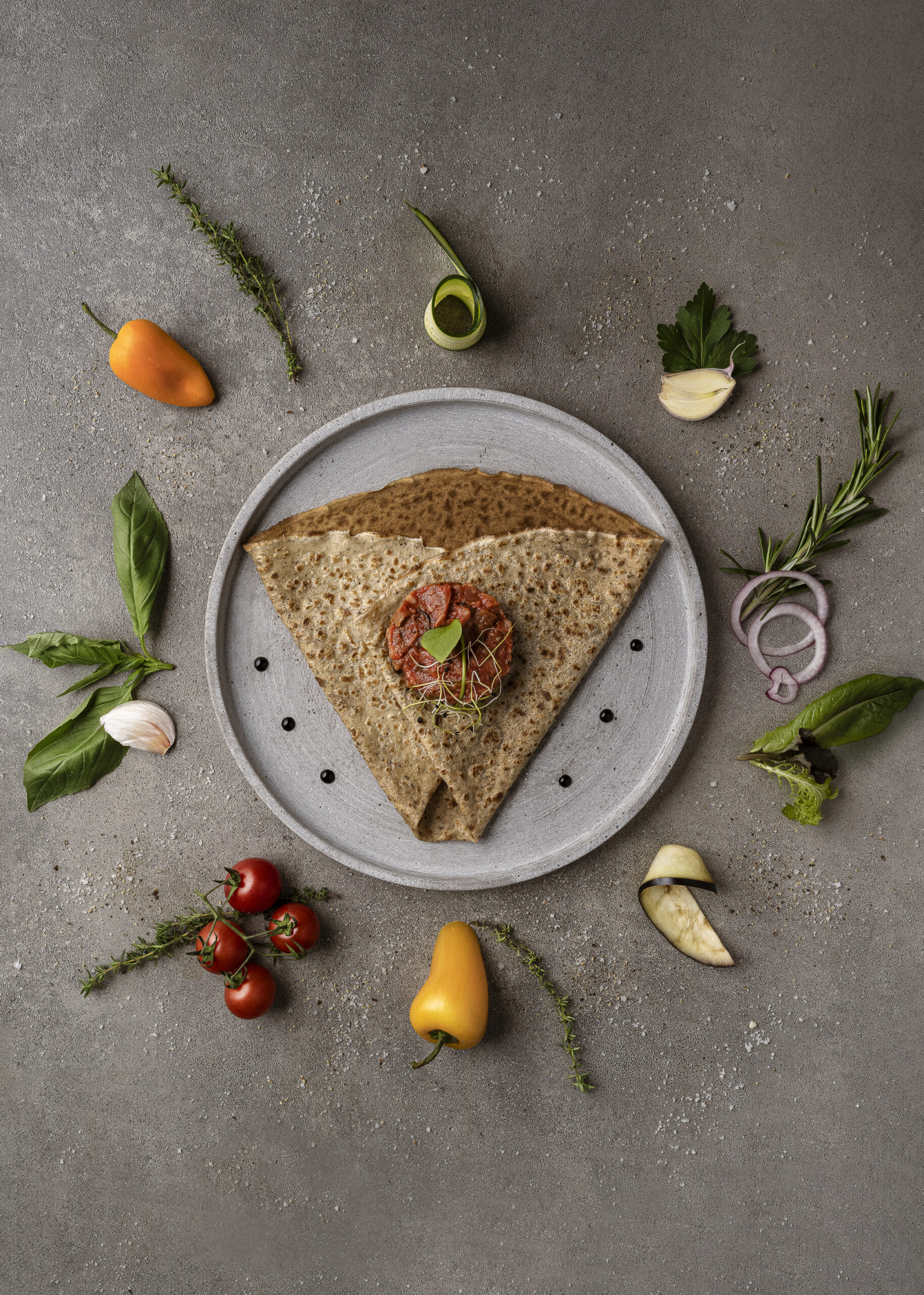 Photographie culinaire, réalisée en studio photo professionnel à Neuchâtel en Suisse. Crêpe vue de dessus sur un fonf béton ciré gris clair avec des ingrédients posés tout autour de la crêpe qui viennent et apportent la création de l'image. Foodphotography shooting photo par bacchus production visuelle. crêpe maison, réalisé avec amour avec plein de légume frais de la ferme.